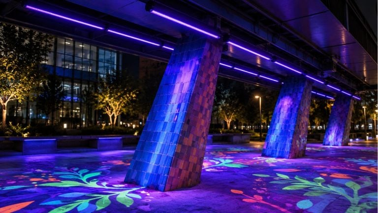 Blacklight fixtures illuminating fluorescent artwork beneath an outdoor building canopy at night.